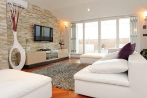 Contemporary living room with a balcony view. There's a white sofa adorned with two oversized white pillows and one small purple decorative pillow. A flat-screen TV is hung on a natural stone wall across the sofa. Two ottomans and a shag rug lie along the center of the room.
